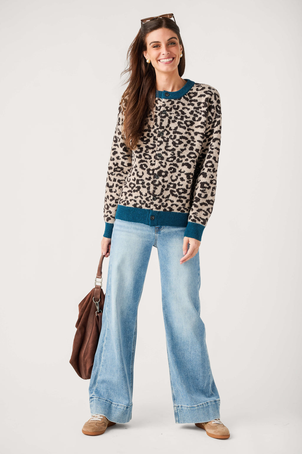 A smiling woman wears a button-front leopard print cardigan with teal trim, wide-leg light jeans, tan sneakers, and carries a brown suede bag.