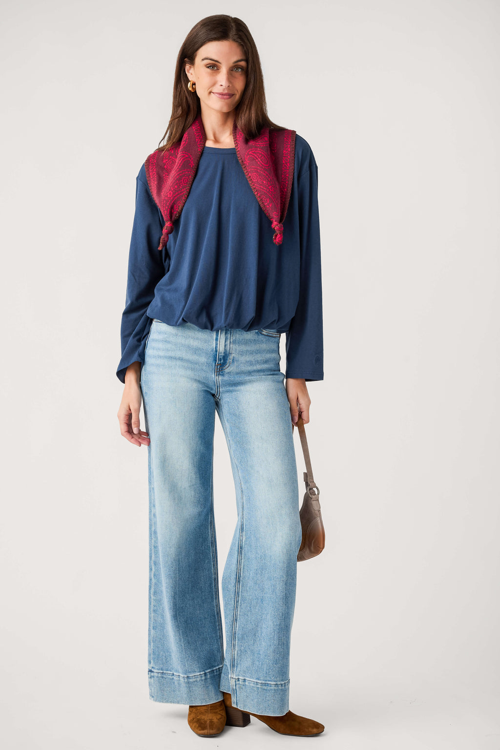 A woman modeling a navy blue long-sleeve top, light-wash wide-leg denim jeans, a dark red scarf, and brown suede boots against a plain light gray studio background.