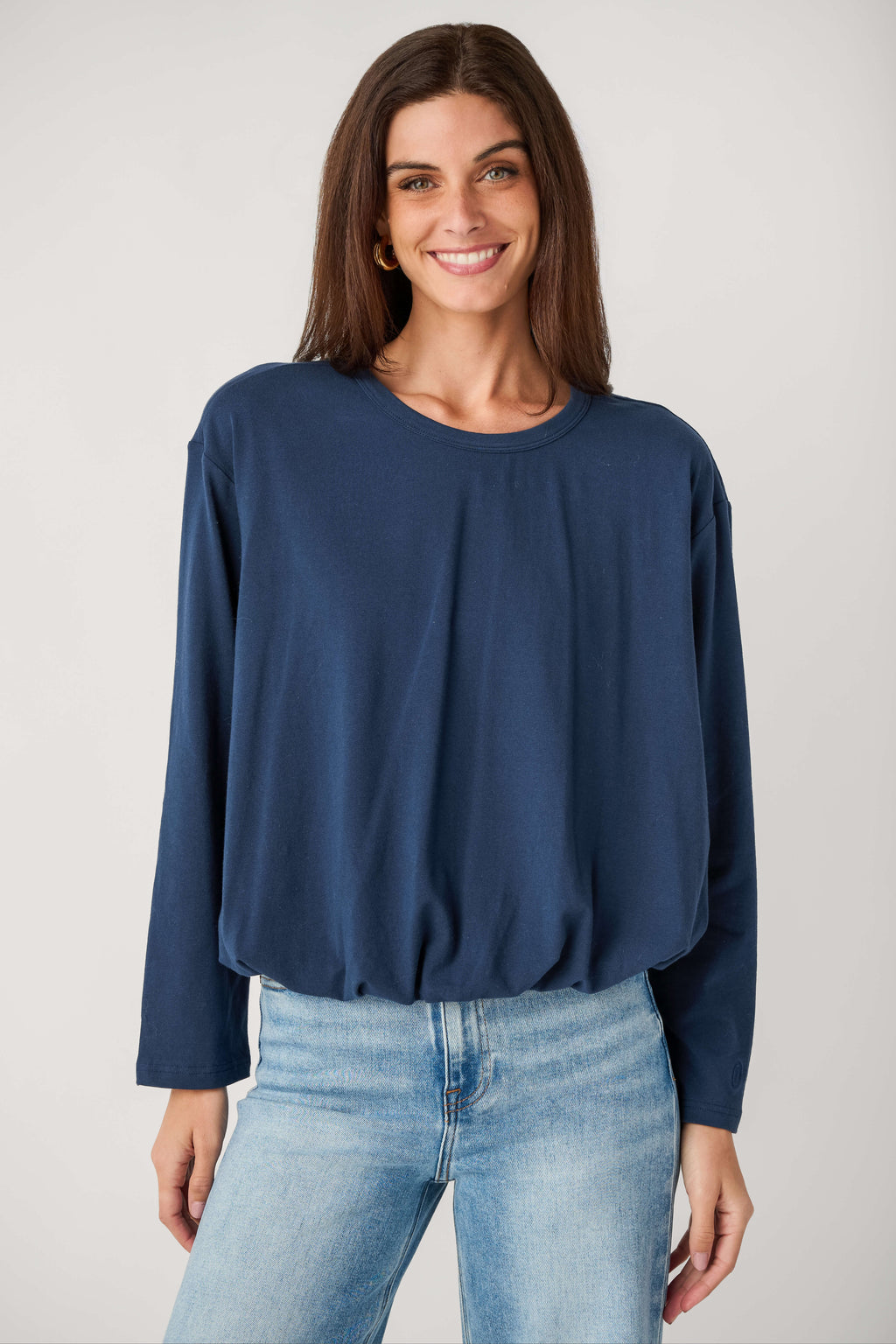 A smiling woman wears a navy long-sleeve crew neck top with a gathered hem, paired with light wash jeans.