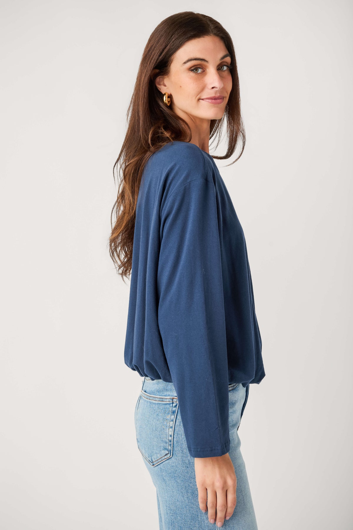 A woman with long brown hair faces sideways wearing a navy long-sleeve top with a gathered hem, light-wash jeans, and gold hoop earrings.