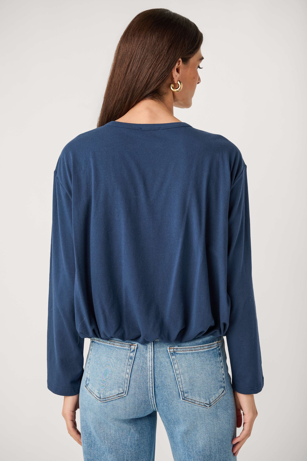 Back view of a woman wearing a navy long-sleeve top with a gathered hem, light-wash jeans, and gold hoop earrings.