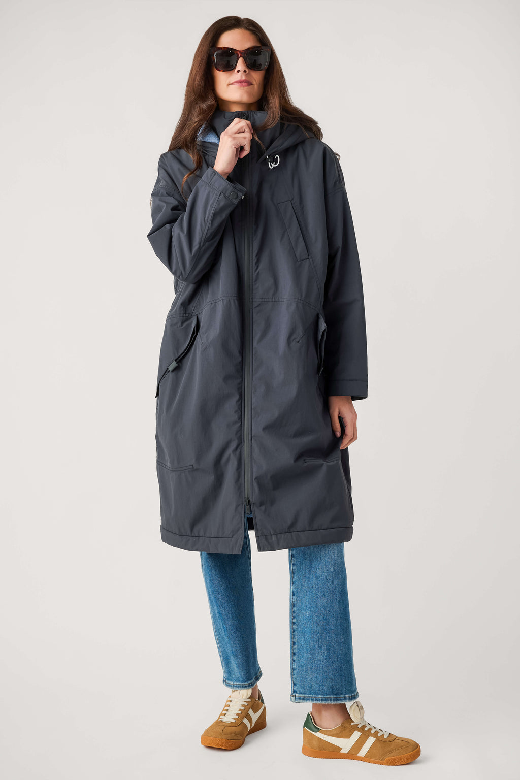 A woman wears a long dark gray hooded parka with a front zipper, blue jeans, tan sneakers, and sunglasses.