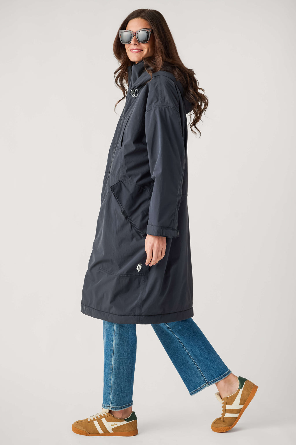 A woman in sunglasses wears a long navy hooded parka with large pockets, blue jeans, and tan suede sneakers.