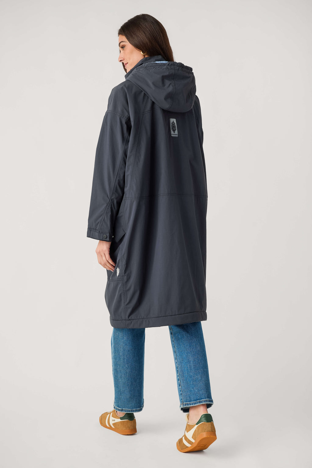 Back view of a woman wearing a long dark navy hooded parka with a small patch, blue jeans, and tan sneakers.
