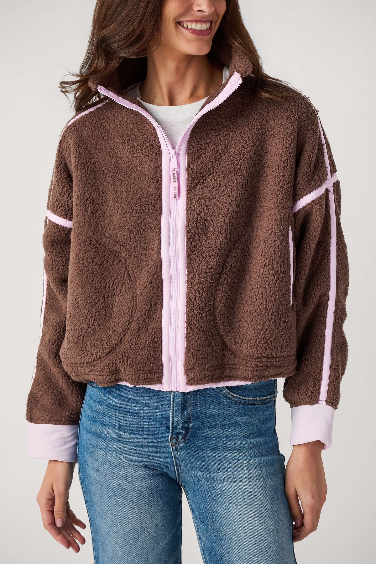 A smiling woman wears a brown sherpa fleece zip-up jacket with light pink trim, a white top, and blue jeans.