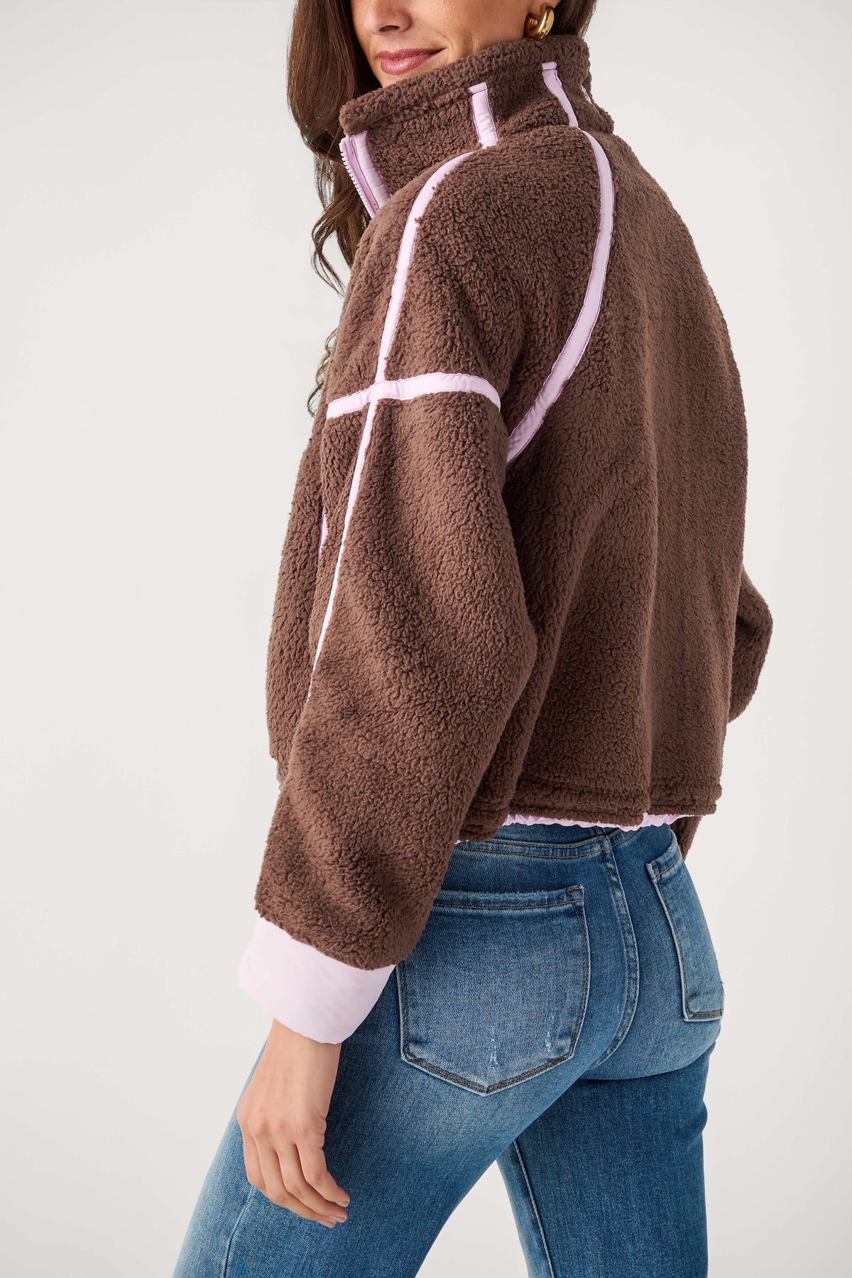 A woman wears a cropped brown sherpa fleece jacket with light pink piping and cuffs, paired with blue jeans, viewed from behind.