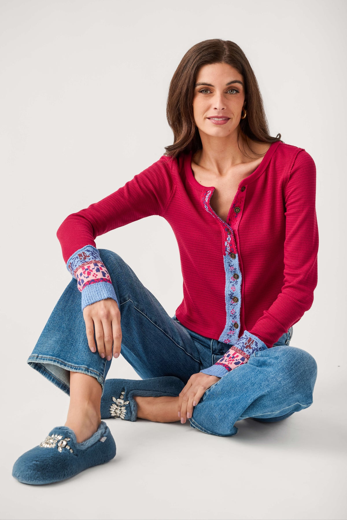 A seated woman wears a red waffle-knit button-front cardigan with blue patterned trim and cuffs, paired with jeans and blue slippers.
