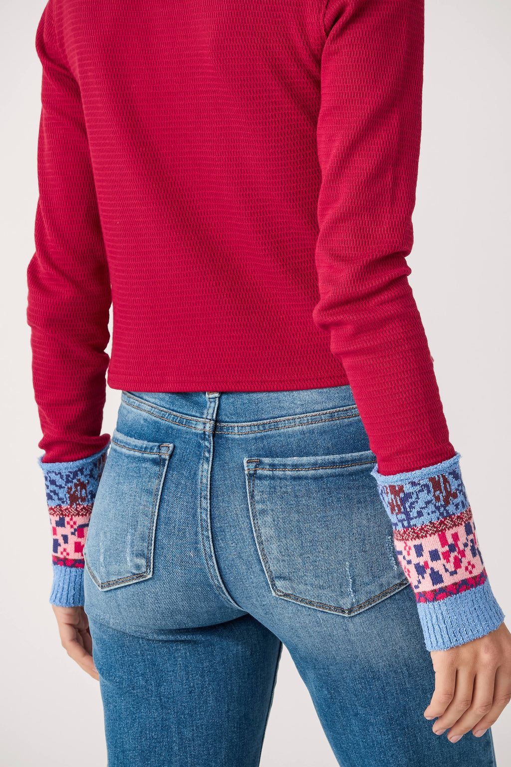 Back view of a red textured long-sleeve top with blue and pink patterned knit cuffs, worn with medium-wash denim jeans.