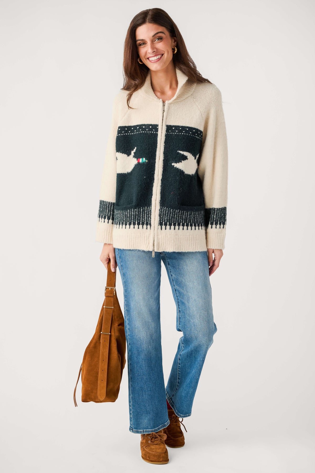 A woman modeling a cream and navy knit zip-up cardigan with bird motifs, paired with blue jeans, brown boots, and a brown tote bag against a light gray studio background.