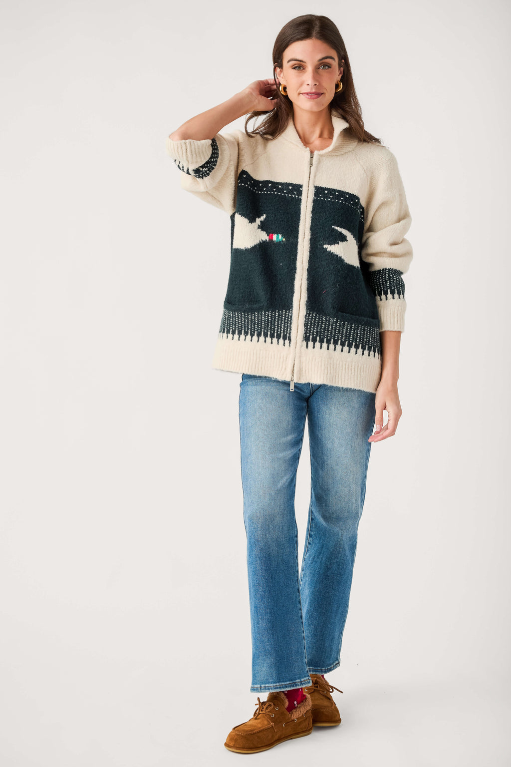 A woman wears a cream zip-up cardigan with a dark green bird pattern, blue jeans, and brown suede shoes.