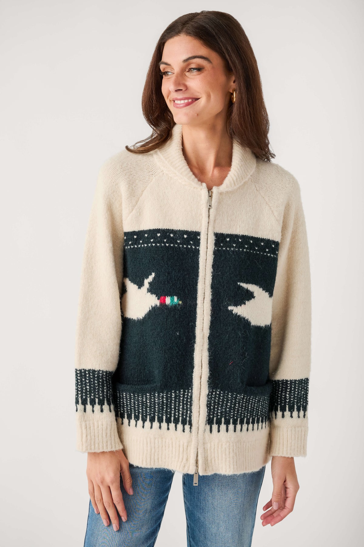A smiling woman wears a cream zip-front shawl collar cardigan with a dark green bird pattern panel, paired with blue jeans.