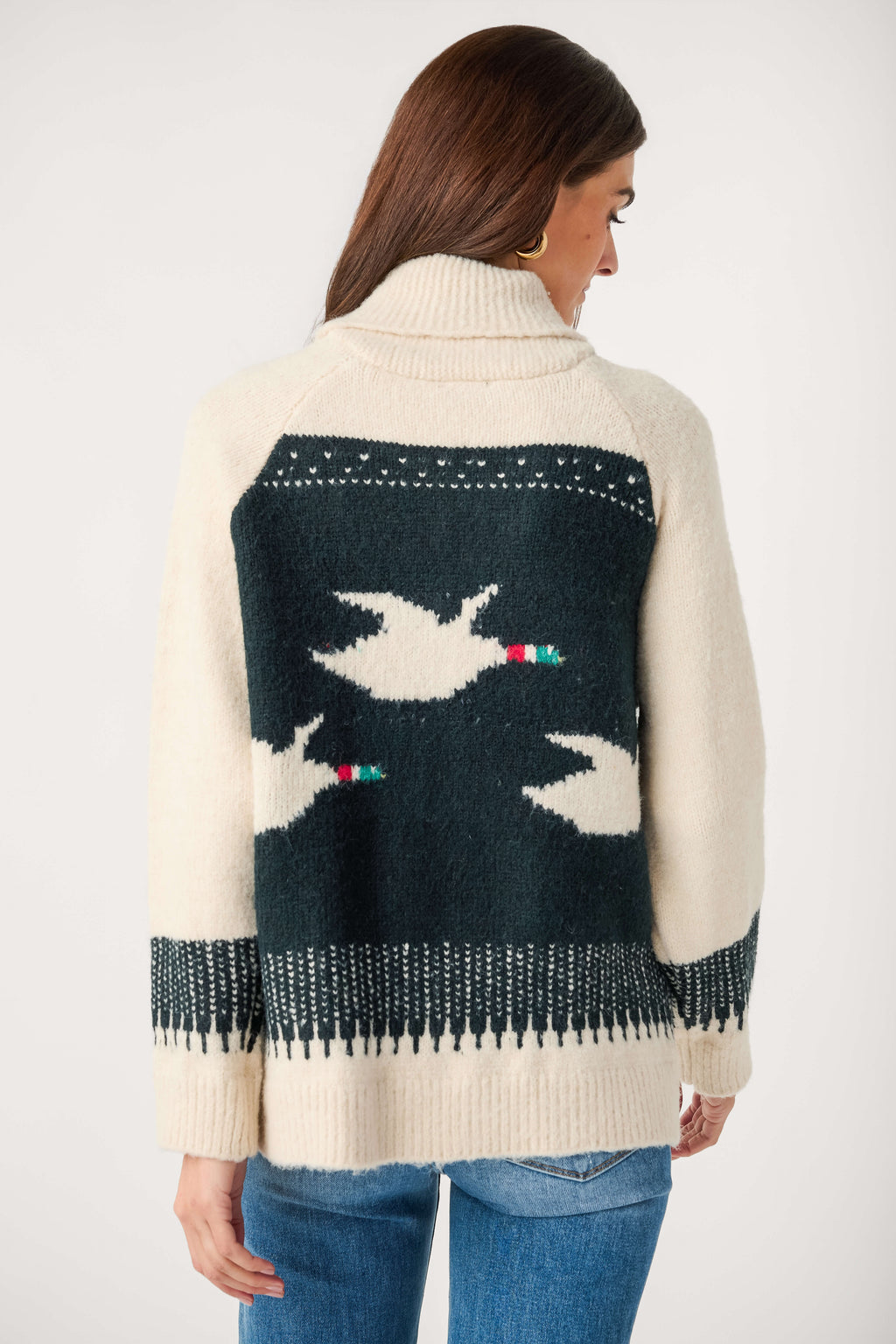 A woman seen from behind wears a cream knit cardigan with a dark green back panel showing white flying birds, paired with jeans.