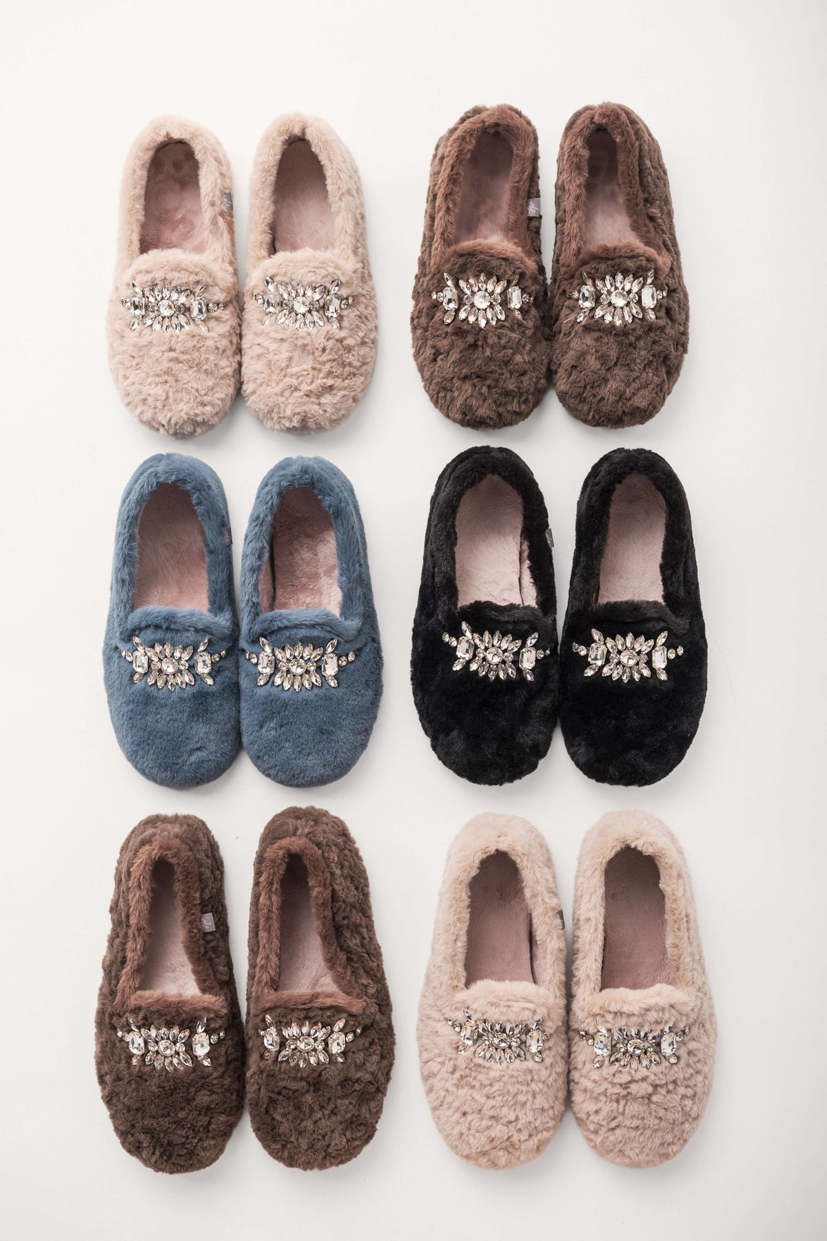 Six pairs of fuzzy loafer slippers in beige, brown, blue, and black, each decorated with a crystal embellishment, arranged in rows.
