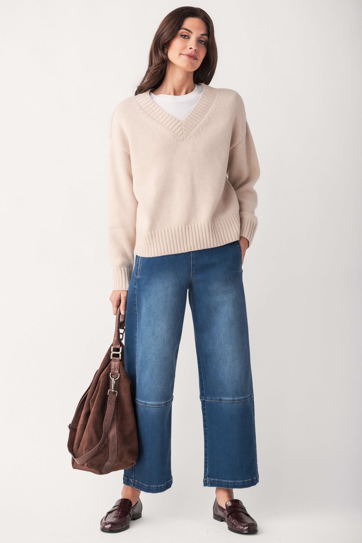 A woman wears a cream ribbed V-neck sweater over a white top with wide-leg jeans, holding a brown suede bag.