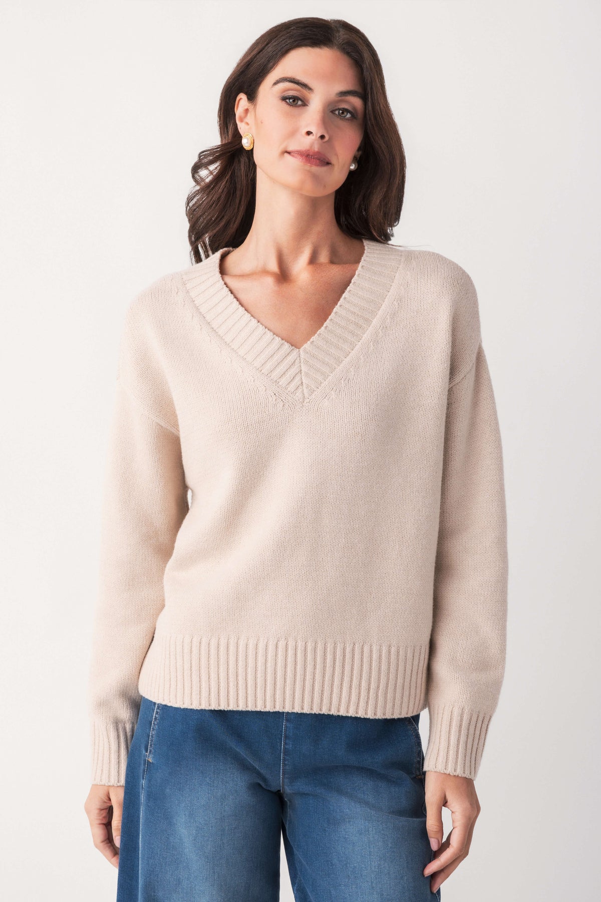 A woman with wavy dark hair wears a cream V-neck sweater with ribbed trim, paired with wide-leg blue jeans.