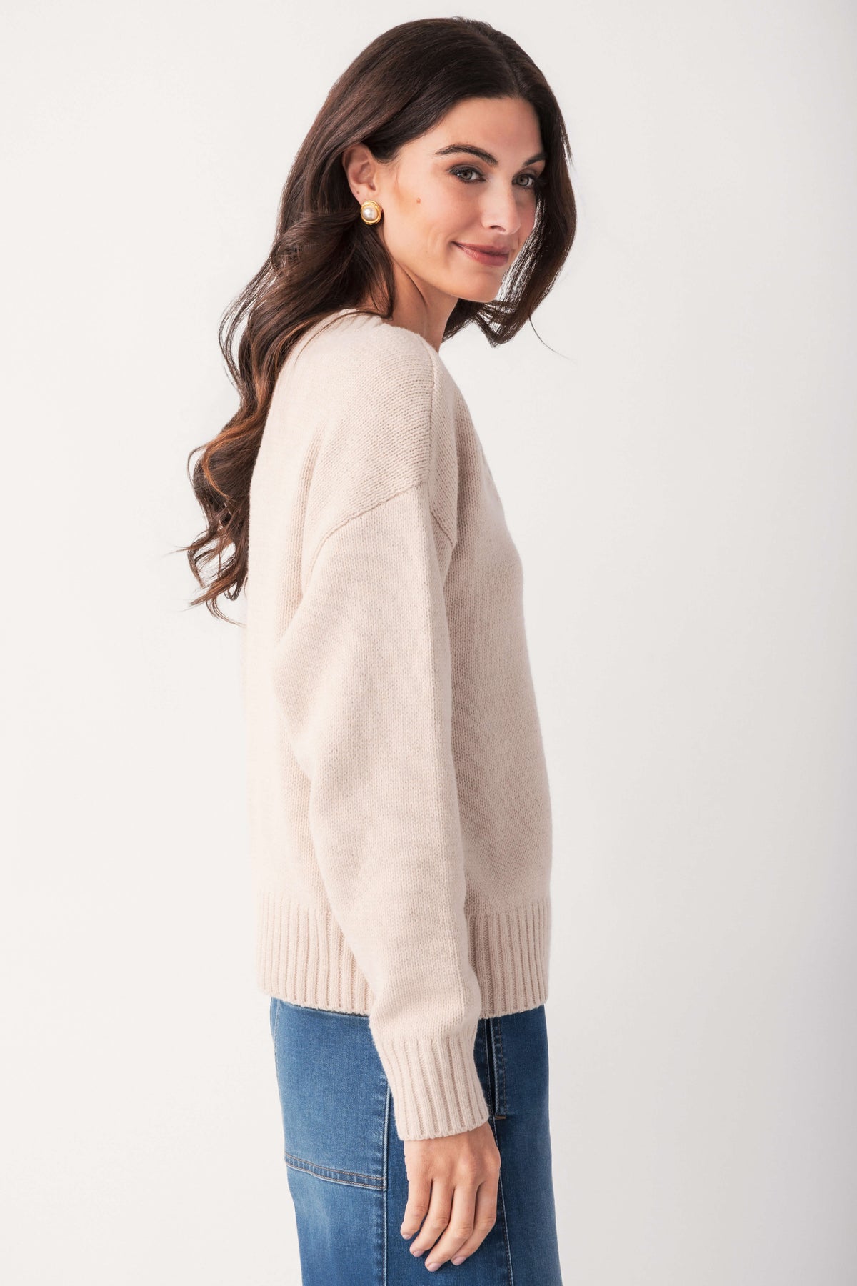 A woman with long dark wavy hair models a cream knit sweater with ribbed cuffs and hem, paired with blue jeans.