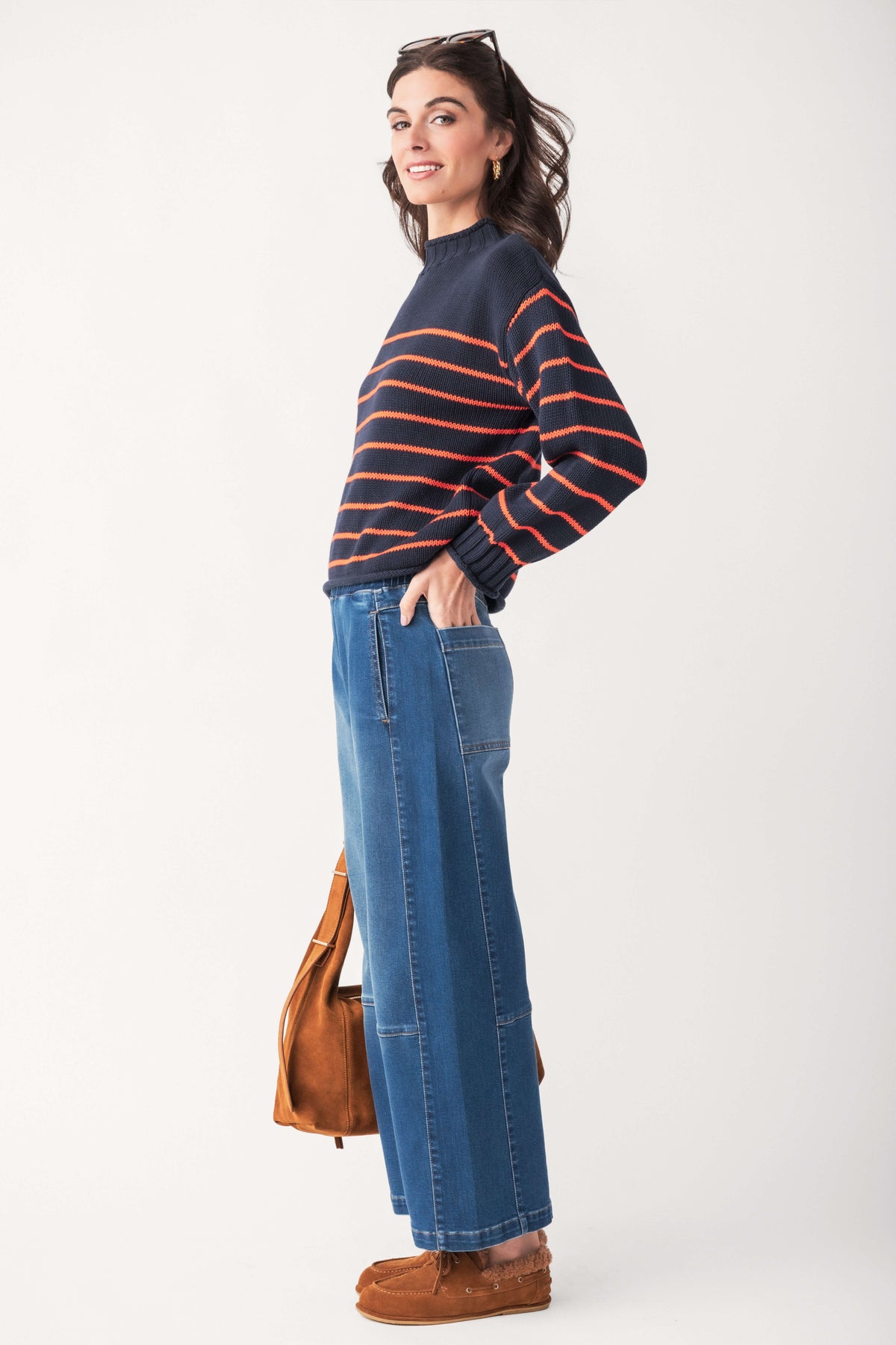A woman in a navy mockneck sweater with orange stripes, wide-leg blue jeans, and brown suede shoes holds a matching suede bag.