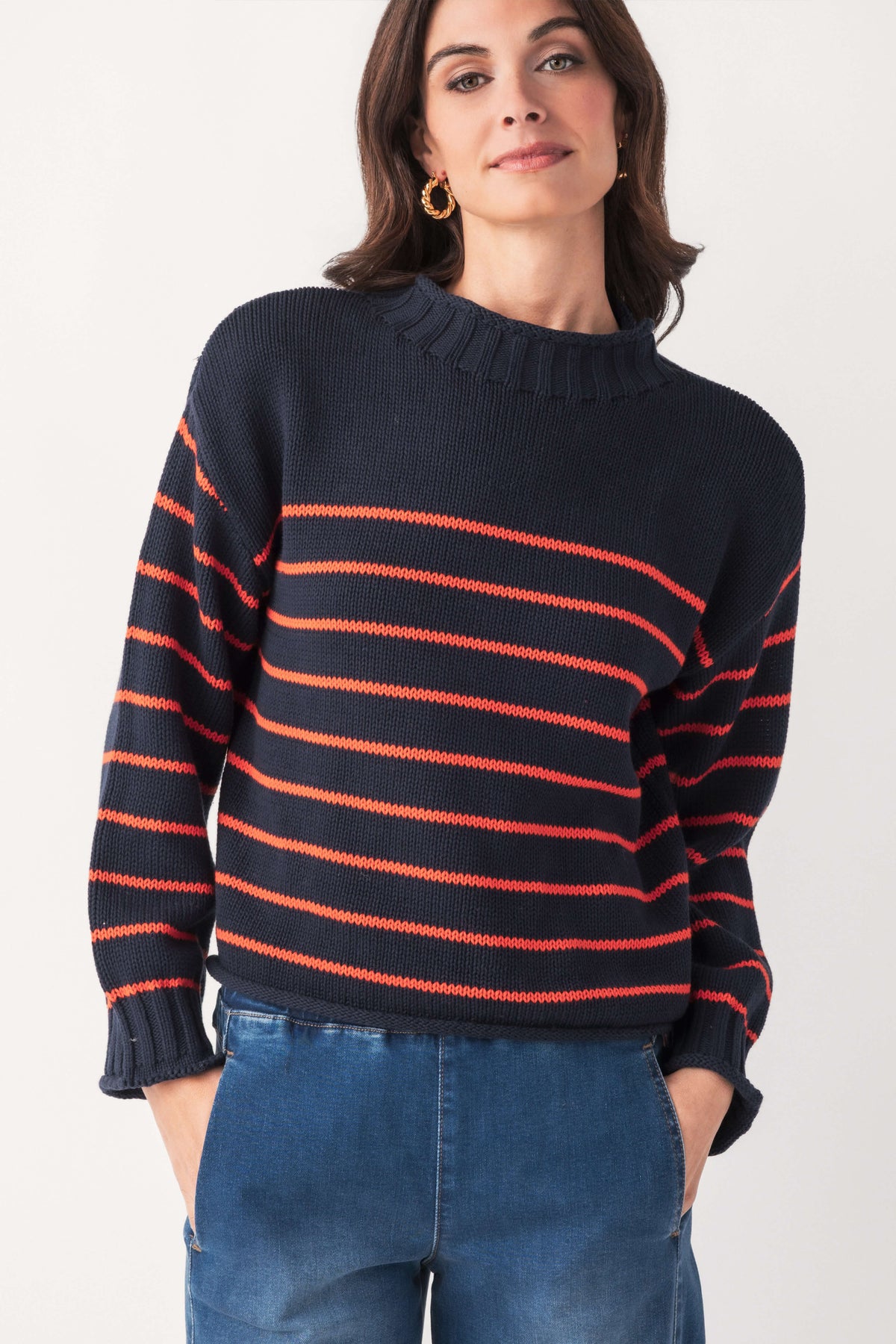 A woman wears a navy knit mockneck sweater with thin coral stripes, paired with blue jeans and gold hoop earrings.