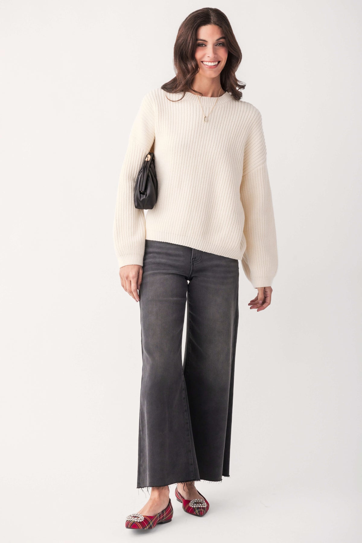 A smiling woman wears a cream ribbed knit pullover with gray wide-leg jeans, red plaid flats, and a small black handbag.