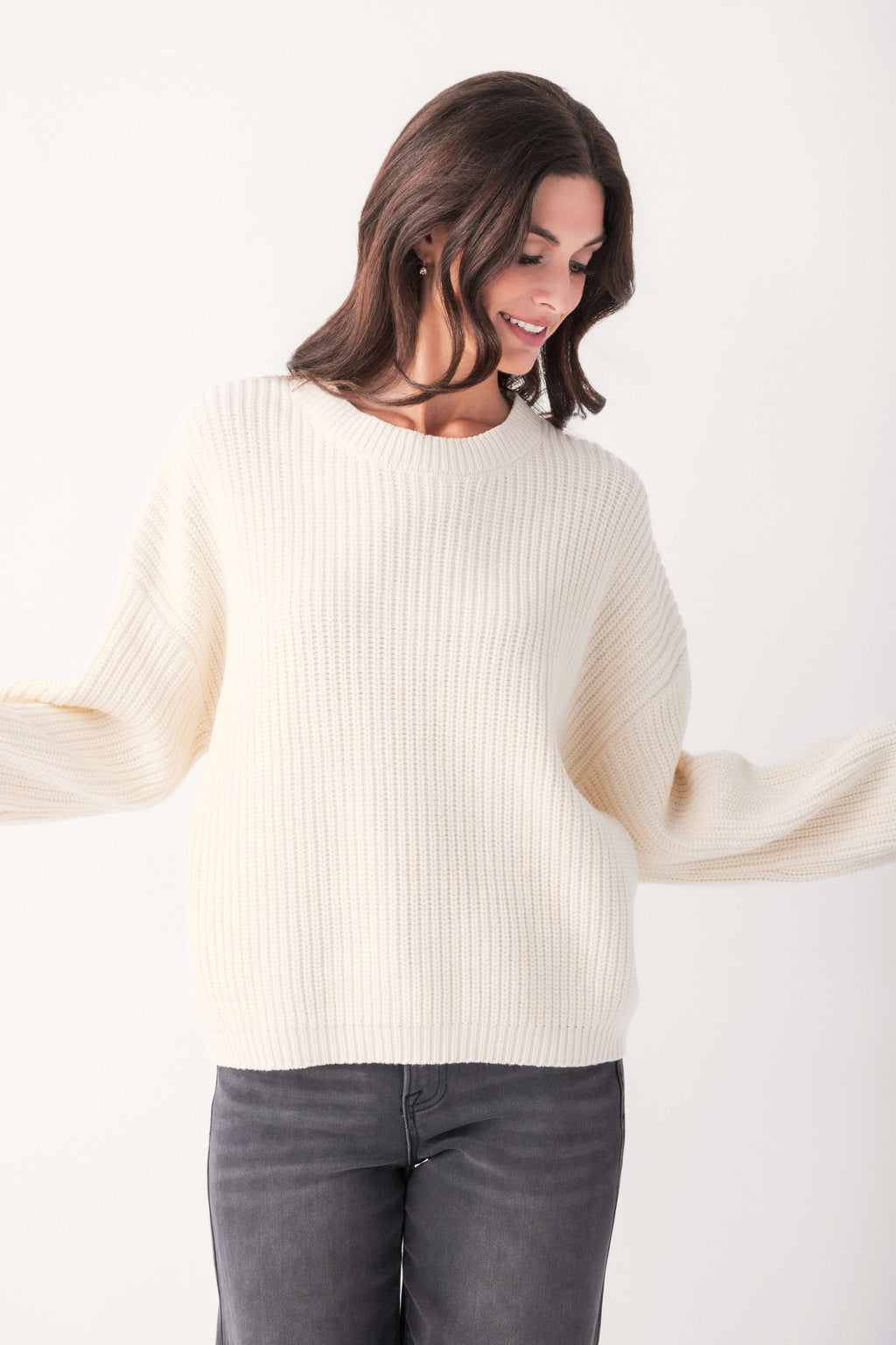 A smiling woman with dark wavy hair wears a cream ribbed knit crewneck sweater with wide sleeves and gray jeans.