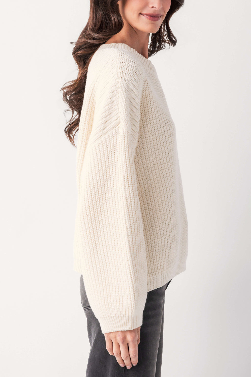 A woman in profile wears a cream chunky ribbed knit pullover with long sleeves and gray jeans against a plain background.