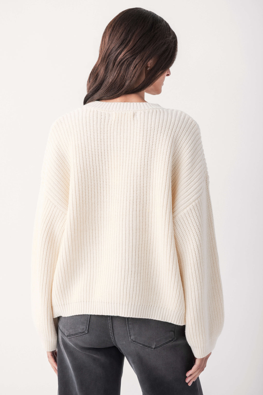 Back view of a woman with dark hair wearing a cream ribbed knit sweater with dropped shoulders and gray jeans.