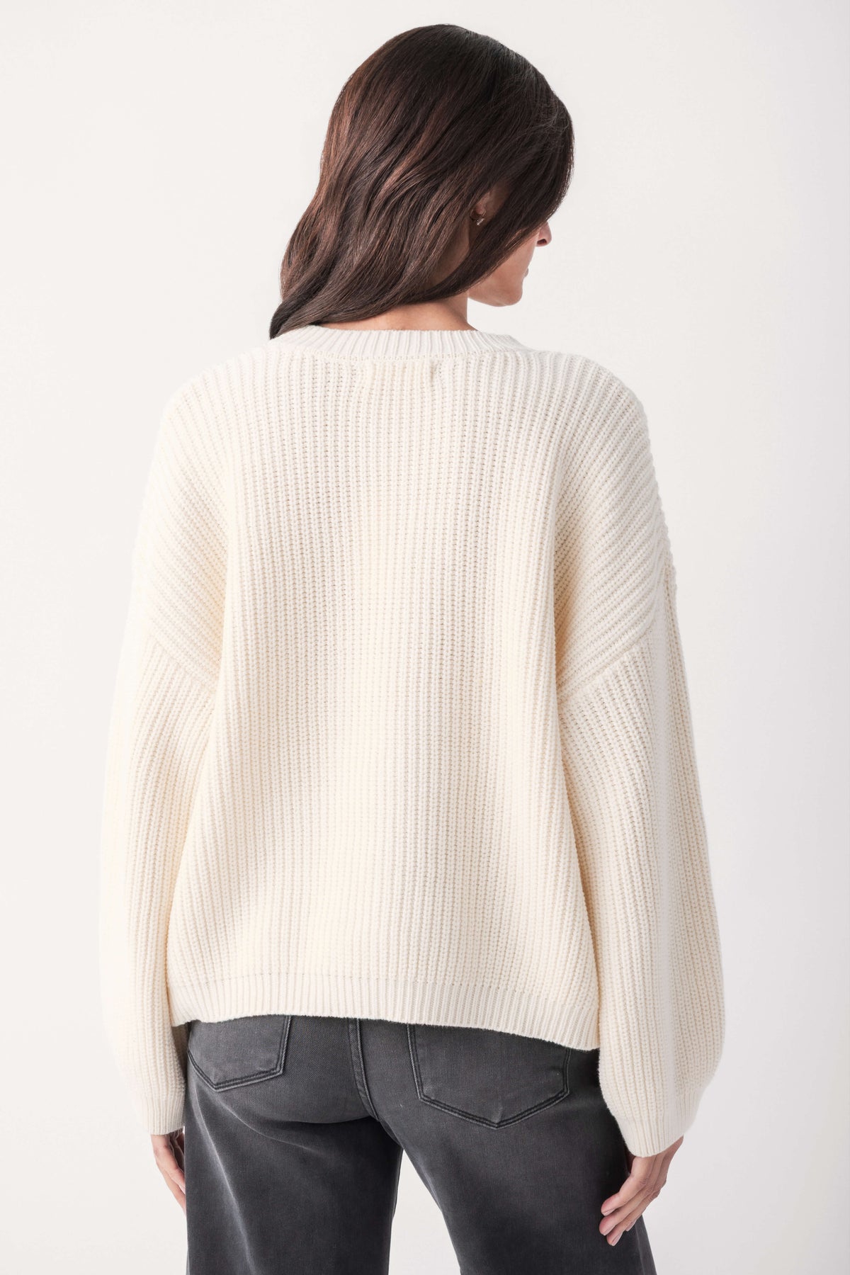 Back view of a woman with dark hair wearing a cream ribbed knit sweater with dropped shoulders and gray jeans.